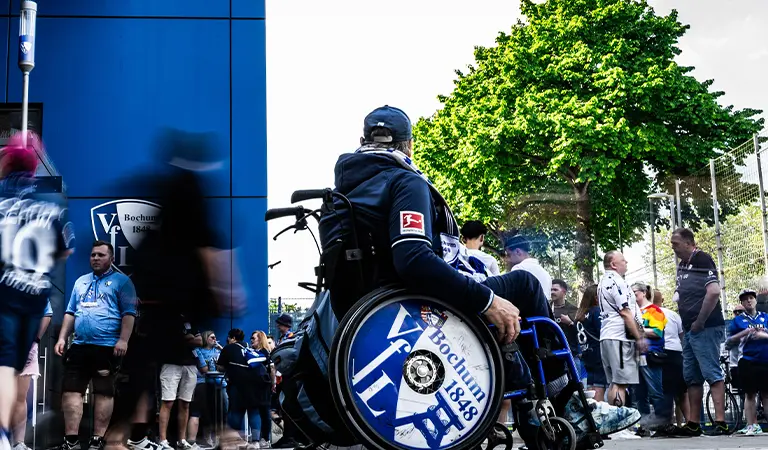 Ein Mann im Rollstuhl im Fokus mit dem VfL Bochums Mannschaftslogo auf diesem Rad als Dekoration, in der Menge der Fans vor dem Stadion. (Foto)
