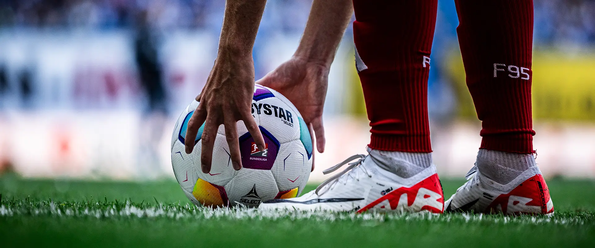 Eine Nahaufnahme eines Fußballspielers, der den Ball für einen Anstoß liest, wobei seine Hände am Ball sichtbar sind und der grüne Rasen unter ihm. (Foto)