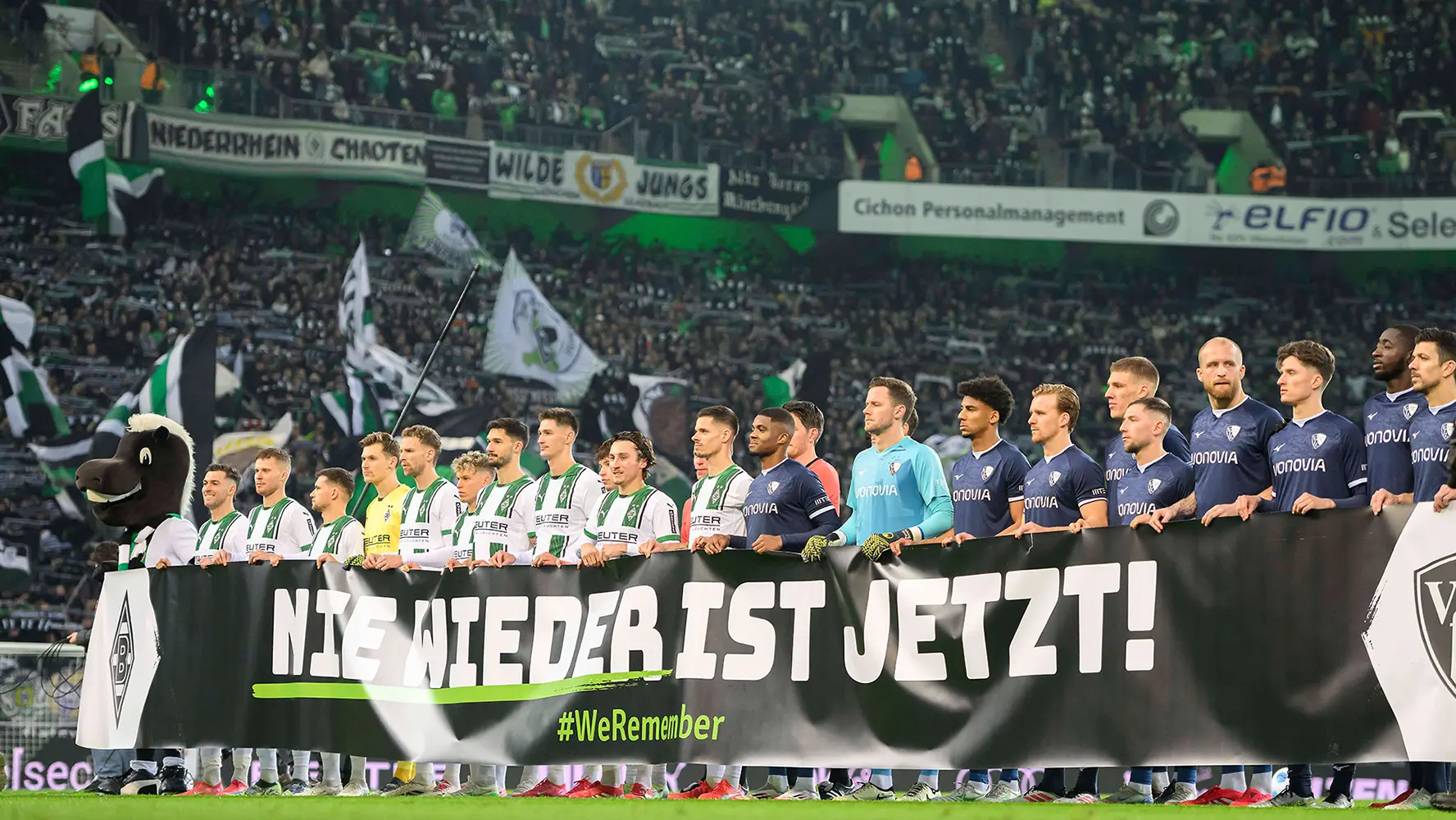 Beide Mannschaften des 19. Spieltags von Borussia Mönchengladbach und VfL Bochum halten ein „Nie wieder ist jetzt!“-Banner auf dem Spielfeld hoch. (Foto)
