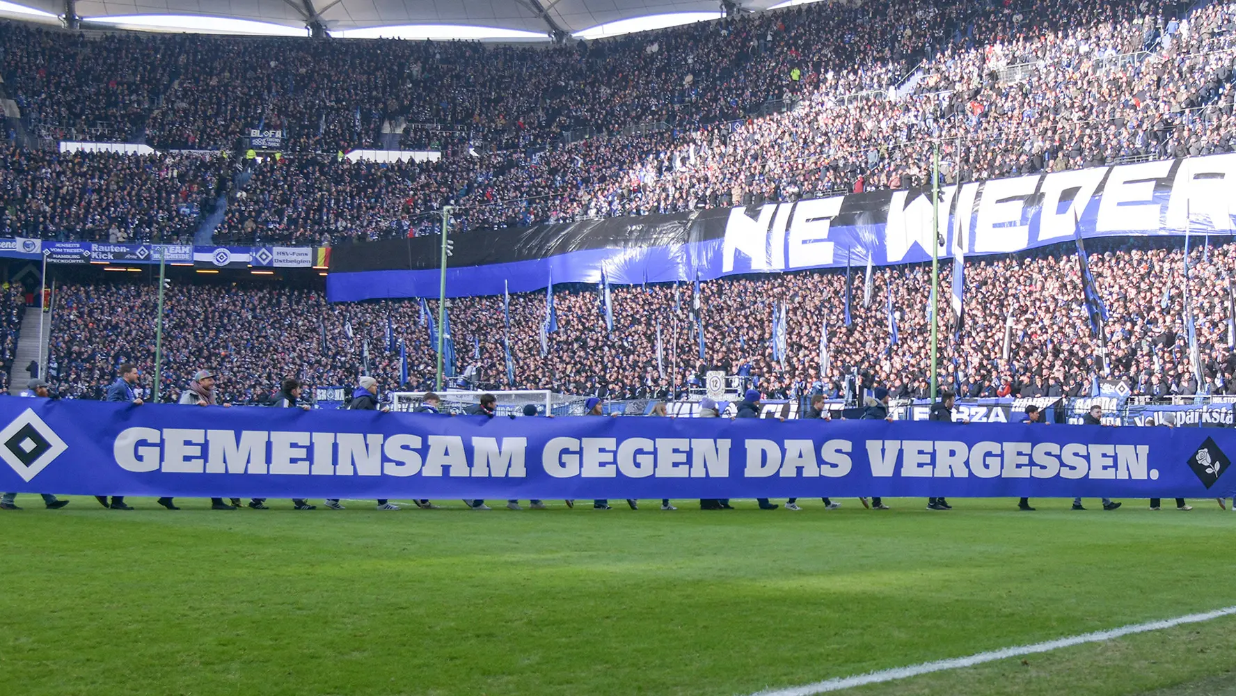 Teammitglieder beider Mannschaften tragen ein großes Banner mit "Gemeinsam gegen das Vergessen" auf das Spielfeld. (Foto)