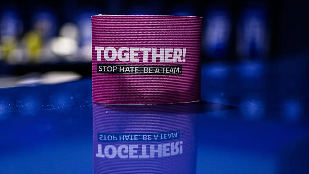 Eine Armbinde mit der Aufschrift "TOGETHER! STOP HATE. BE A TEAM."-Logo auf blauem, spiegelndem Untergrund. (Foto)