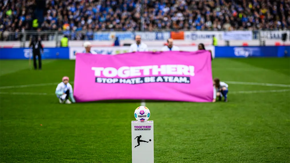 Ein Fußball auf einem Podest mit der Aufschrift "TOGETHER! STOP HATE. BE A TEAM."-Logo und einige Teammitglieder, die ein lilafarbenes Banner mit demselben Logo im unscharfen Hintergrund hochhalten. (Foto)