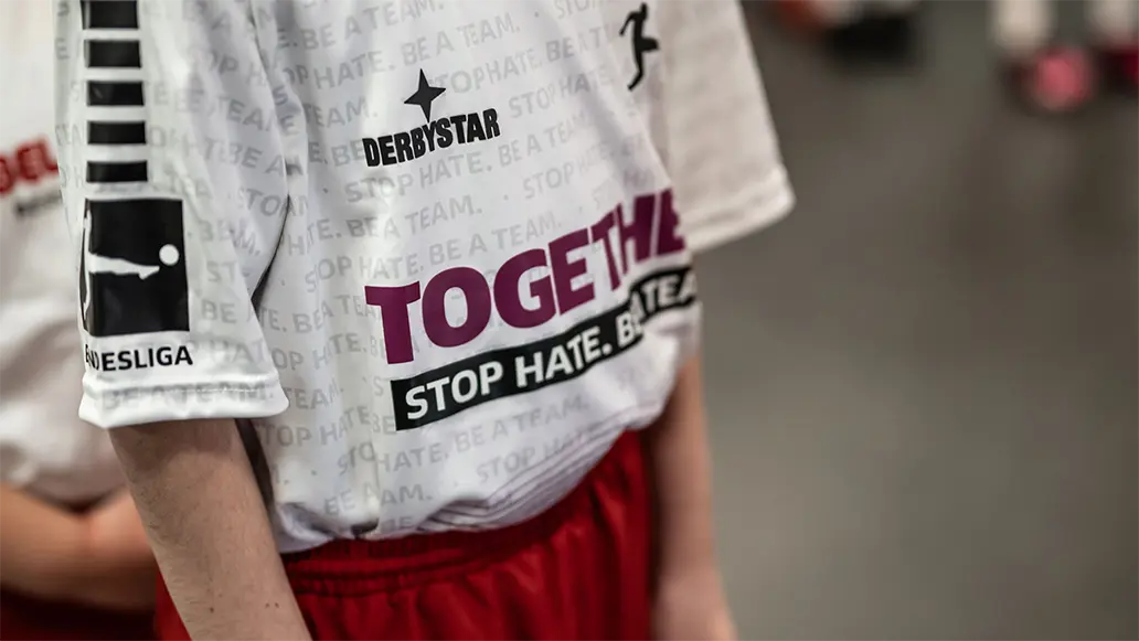 Nahaufnahme eines Kindes, das ein weißes Trikot mit der Aufschrift "TOGETHER! STOP HATE. BE A TEAM."-Logo darauf. (Foto)