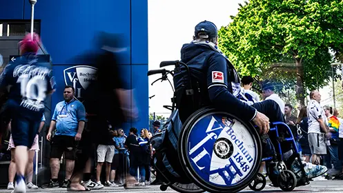 Ein Mann im Rollstuhl im Fokus mit dem VfL Bochums Mannschaftslogo auf diesem Rad als Dekoration, in der Menge der Fans vor dem Stadion. (Foto)