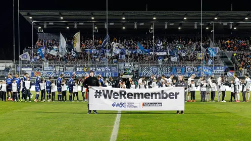 Zwei Personen, die ein Banner mit der Aufschrift #WeRemember hochhalten, mit Spielern und Kindern auf dem Spielfeld im Hintergrund. (Foto)