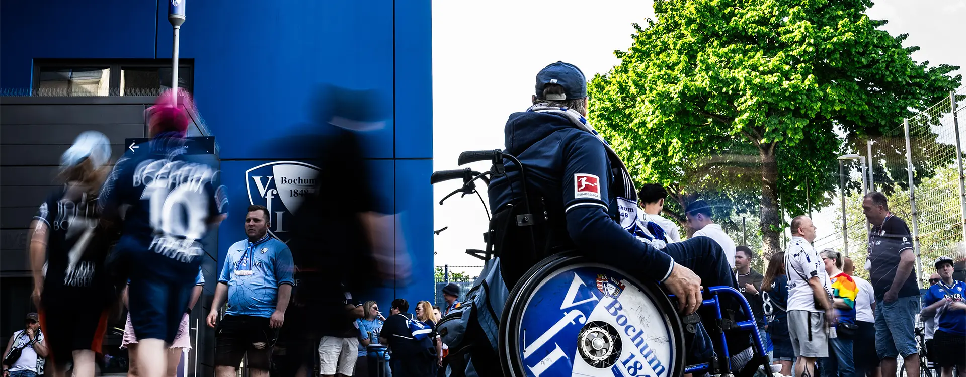A man in a wheelchair in focus with the VfL Bochums team logo on this wheels as decoration, in the crowd of fans before the stadium. (Photo)