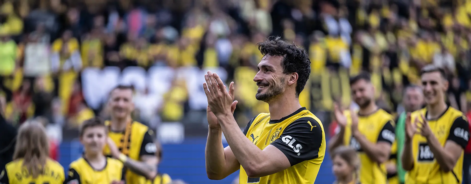 Mats Hummels clapping in his black and yellow jersey owith team mates and fans visible in the background. (Photo)