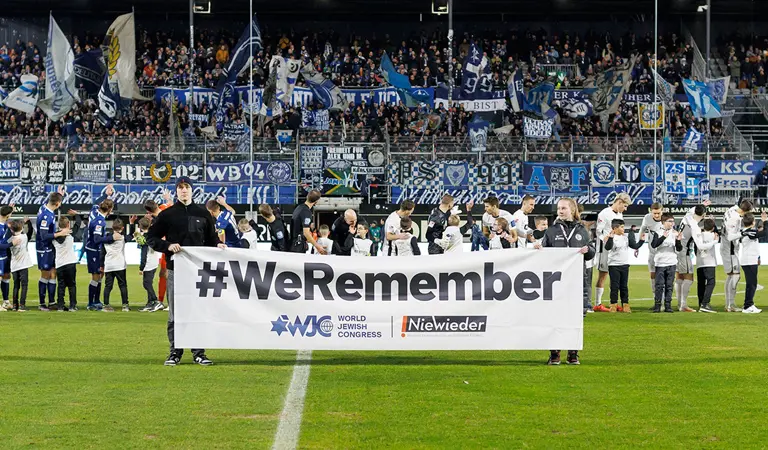 Two persons holding up a banner with #WeRemember, with players and kids on the field in the background. (Photo)