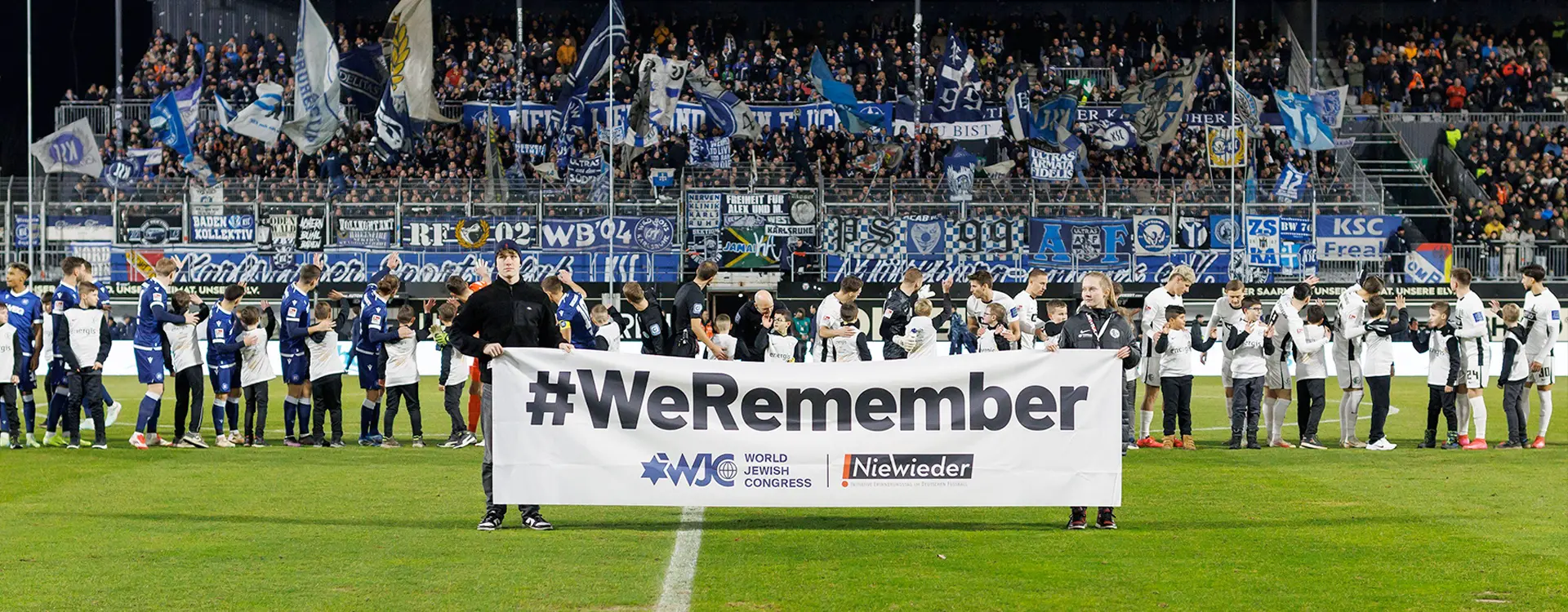 Two persons holding up a banner with #WeRemember, with players and kids on the field in the background. (Photo)