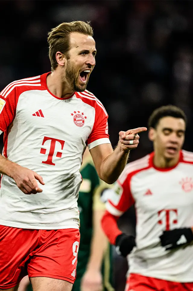 Harry Kane in action in his white red FC Bayern München jersey on the football field, celebrating a goal with a finger wag. (Photo)