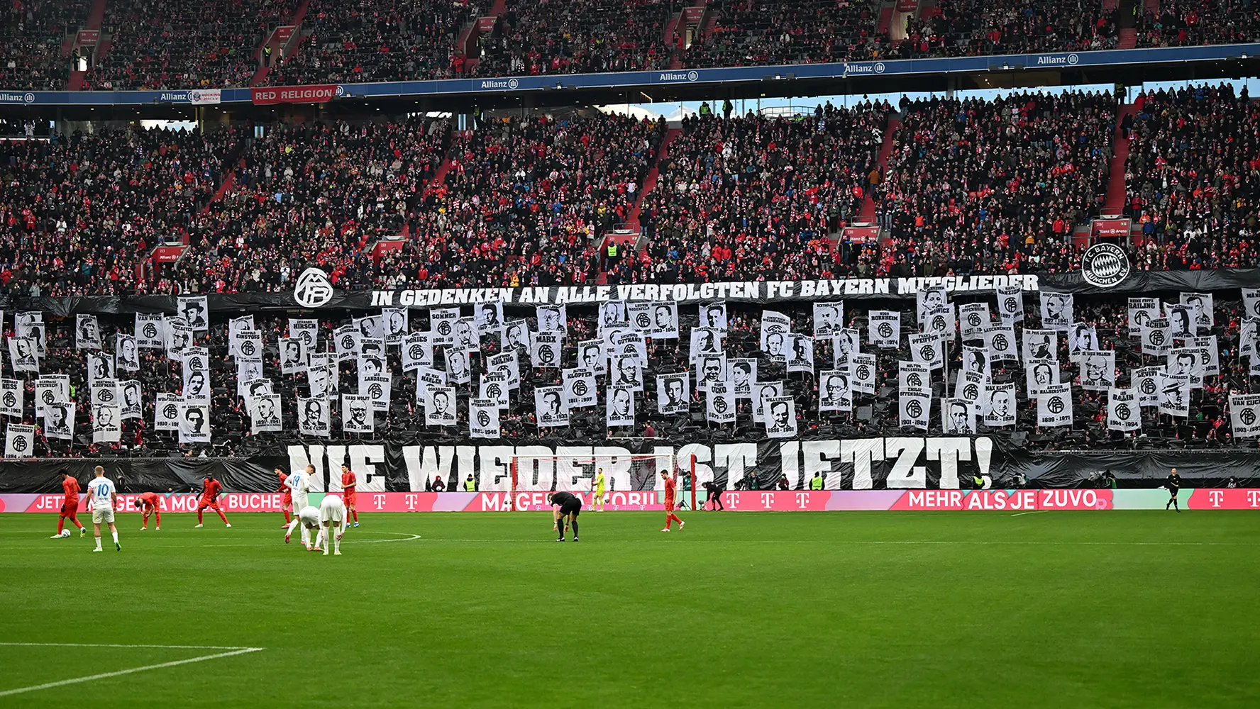 A full stadion with the lower ranks holding up a large "Nie wieder ist jetzt!" banner, with individual profile posters held up behind it. (Photo)