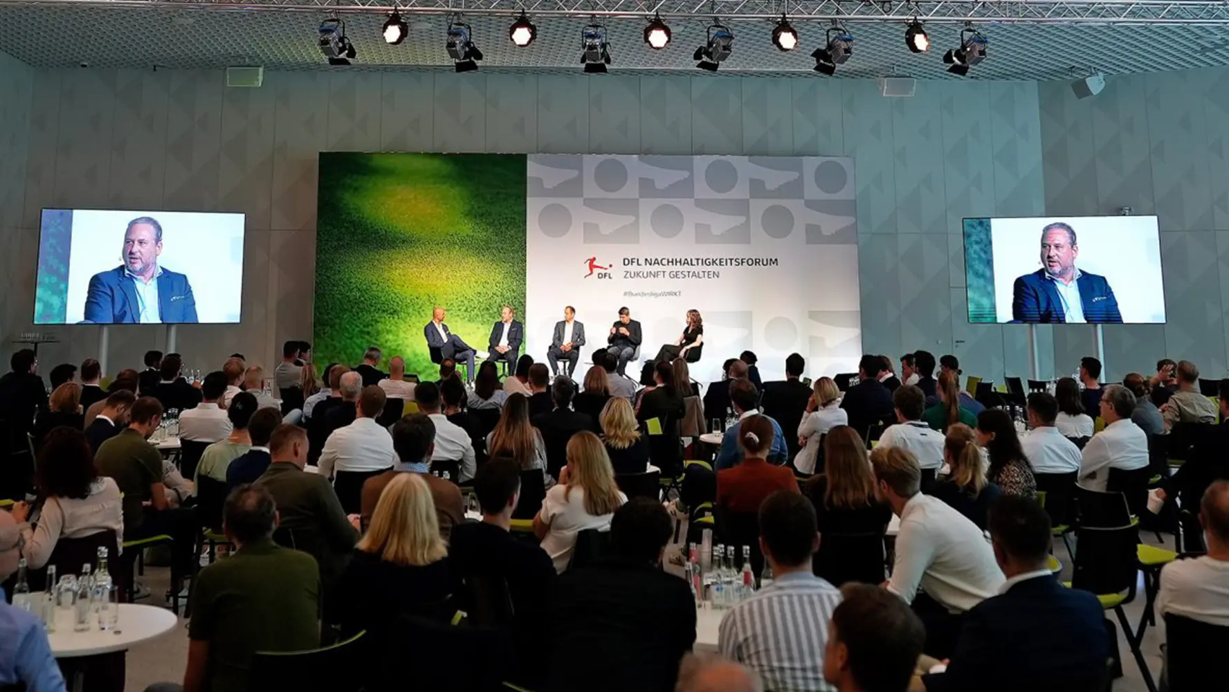 A woman and four men sitting and talking up fron on the stage, and the audience watching them from the floor on round tables. (Photo)