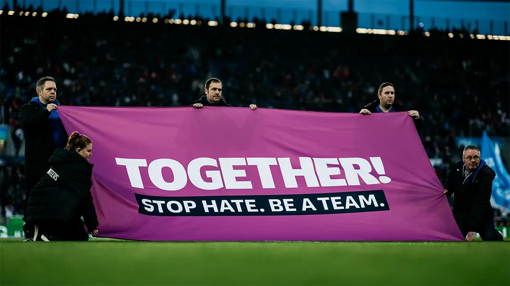 Teamsters hold up a large purple "TOGETHER! STOP HATE. BE A TEAM." banner on the field during pre-game. (Photo)