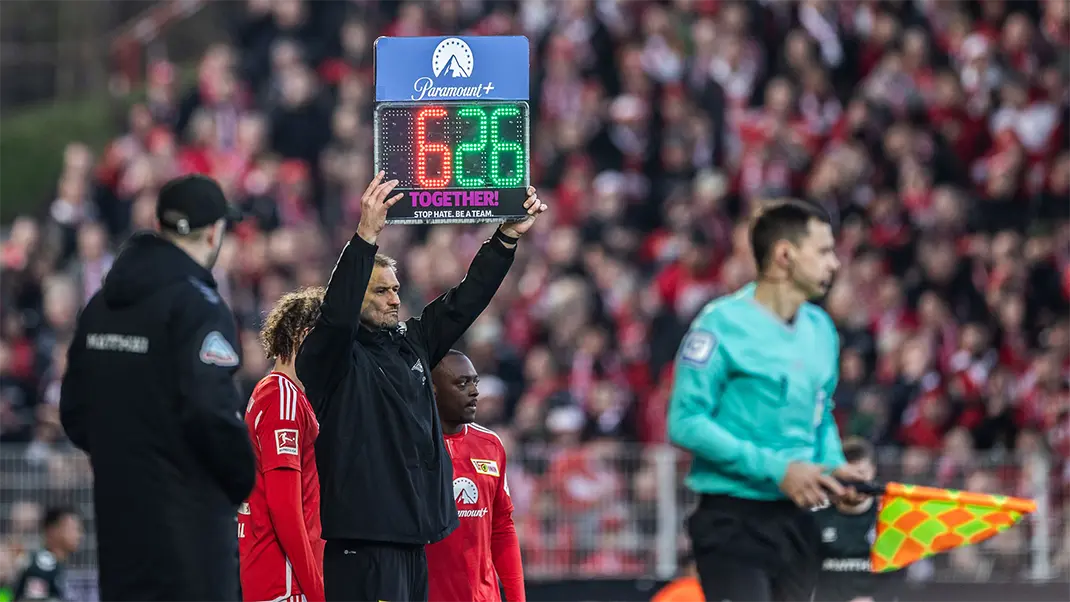 A referee on the sidelines holding up a sign panel for a player exchange and the "TOGETHER! STOP HATE. BE A TEAM." logo on it. (Photo)