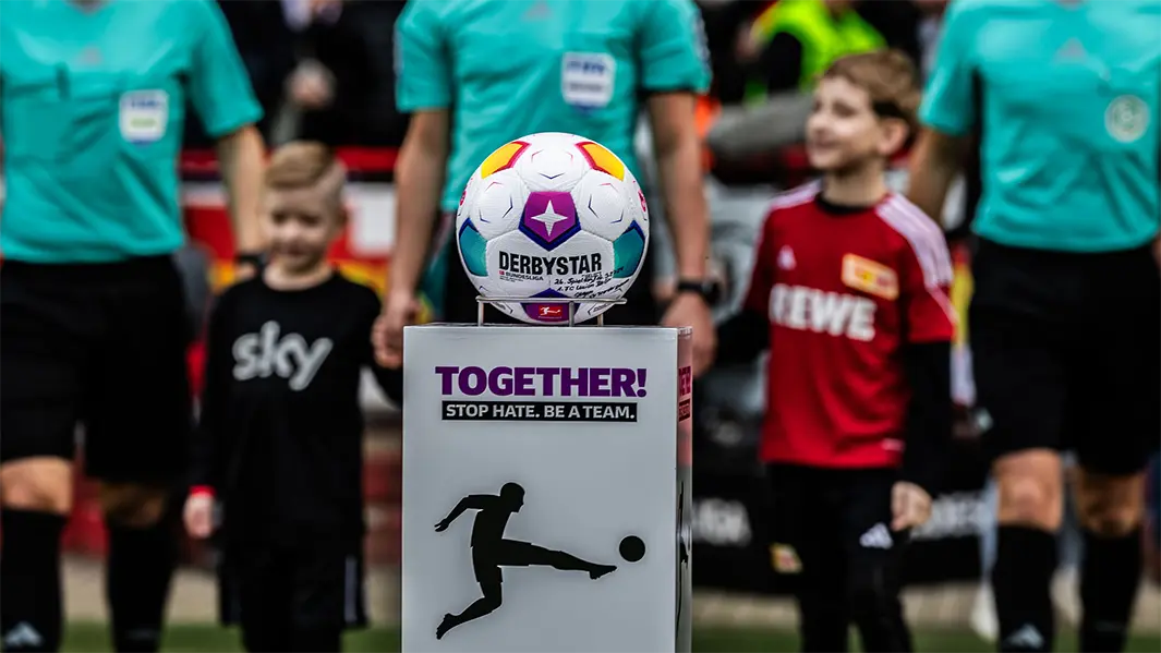 A football placed on a podest with the "TOGETHER! STOP HATE. BE A TEAM." logo, and some football players holding hands with kids in the fuzzy background. (Photo)