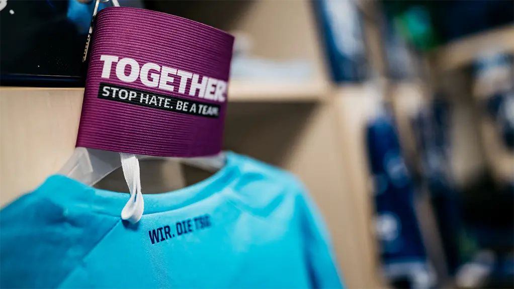 Closeup of an armband with the "TOGETHER! STOP HATE. BE A TEAM." and a jersey on a hanger in the changing room. (Photo)