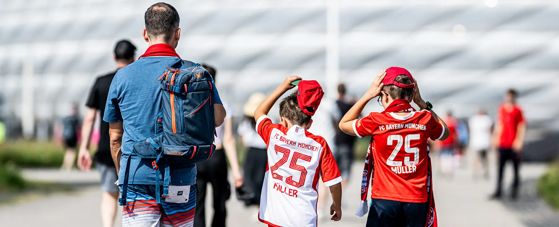 A&nbsp; father in a blue shirt and a blue backpack walks towards the stadium with his two sons in FC Bayern München jerseys and ball caps. (Photo)
